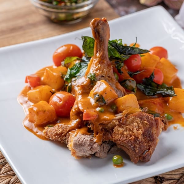Crispy duck leg in red curry sauce with pineapple, cherry tomatoes, and basil on a white plate.
