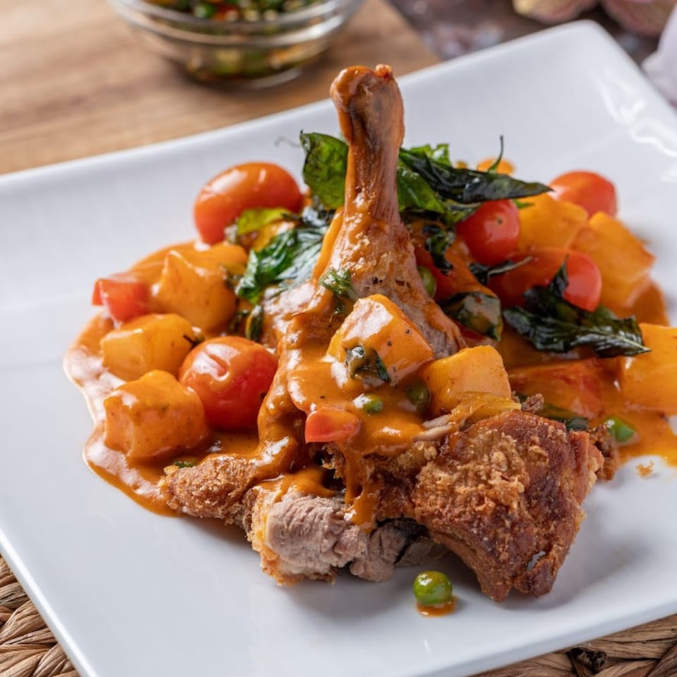Crispy duck leg in red curry sauce with pineapple, cherry tomatoes, and basil on a white plate.