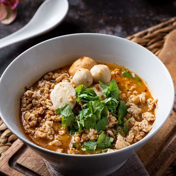 Spicy Thai noodle soup with minced pork, fish balls, and fresh cilantro in a white bowl.