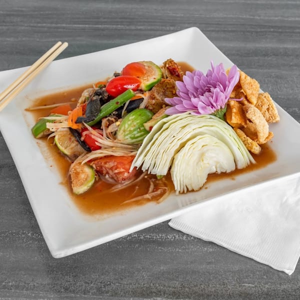 Thai papaya salad with tomatoes, long beans, cabbage, pork rinds, and a purple flower garnish.